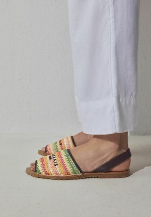 Pieds chaussés de sandales à brides tissées multicolores, associées à un pantalon ample en lin blanc sur un fond clair uni.