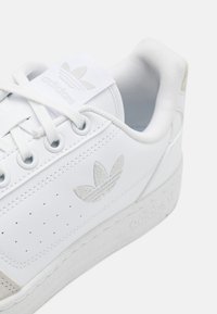 Hvide lædersneakers med perforeret design, teksturerede accenter og logo. Har flade snørebånd og en gummisål med præget branding.