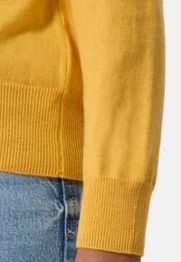 Maglione giallo con polsini e orlo a costine, texture morbida e design aderente, esposto accanto a jeans in denim blu.