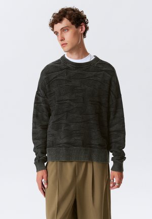 Pull en maille texturé gris foncé avec un motif ondulé, poignets et ourlet côtelés, associé à une chemise blanche en dessous et un pantalon beige ample.