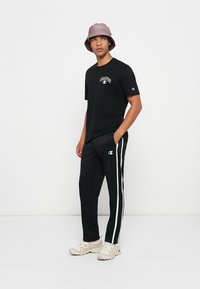 Schwarzes Baumwoll-T-Shirt mit Logo auf der Brust, kombiniert mit schwarzen Jogginghosen mit weißen Seitenstreifen. Warmer Bucket Hat und beigen Sneakern.