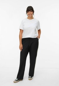 T-shirt blanc à manches courtes, pantalon noir à jambes larges avec détail de bande latérale, baskets grises et vertes, pose debout. Tenue simple et décontractée.