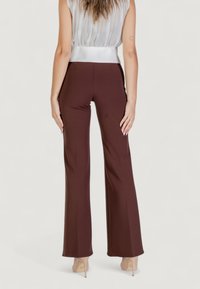 Pantaloni a campana marroni realizzati in tessuto elasticizzato, caratterizzati da una vita alta e una texture liscia, abbinati a scarpe con tacco trasparenti.