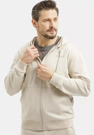 Mężczyzna w beżowej bluzie z zamkiem na szarą koszulką, poprawiający zamek, patrzący lekko w prawo na jednolitym jasnym tle.