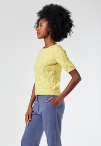 Gelbes kurzärmliges Sweatshirt mit strukturiertem Muster und geripptem Saum, kombiniert mit hellblauen Hosen. Weiches, gestricktes Material mit tailliertem Design.