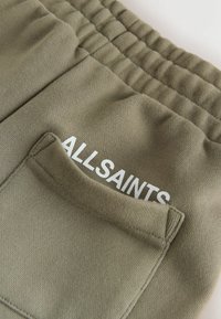 Detail olivovo zeleného odevu s elastickým pásom a vreckom, s čiastočne viditeľným bielym nápisom "ALLSAINTS" nad vreckom.