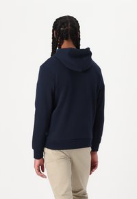 Felpa con cappuccio blu navy fatta di tessuto morbido, con tasca a marsupio e polsini a costine, indossata con pantaloni beige chiaro.