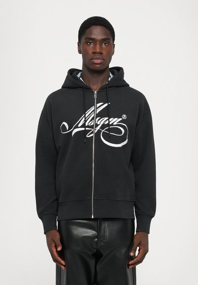 Jeune homme portant un sweat à capuche noir zippé avec un logo en script blanc et un pantalon en cuir noir, se tenant devant un fond blanc uni.