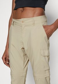 Beige Cargo-Hosen mit schmaler Passform, seitlicher Tasche, Gürtelschlaufen und Knopfverschluss. Aus leichtem Stoff mit einer glatten Textur gefertigt.