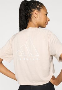 Jasnobeżowa koszulka do biegania z krótkim rękawem, z białym logo "ADIDAS" i napisem "RUNNING" na plecach; luźny fason z rękawami raglanowymi.