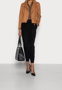 Veste en cuir suède beige, chemise à carreaux noire, joggings noirs, escarpins noirs à talons, et un sac à main noir structuré avec des anses.
