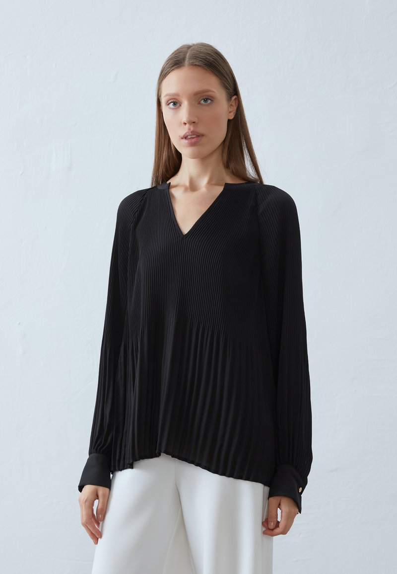 Haut plissé noir avec décolleté en V, manches longues et poignets boutonnés. Fabriqué en tissu léger, avec une silhouette fluide.