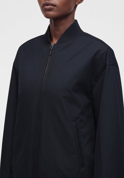 Άνω μέρος σώματος ατόμου που φοράει μαύρο bomber jacket με φερμουάρ, με ριμπ ύφασμα στον γιακά και ορατές πλαϊνές τσέπες, σε απλό φόντο.