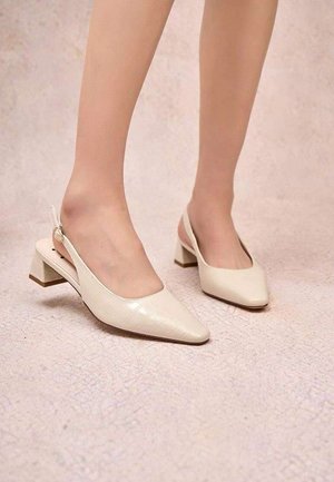 Pieds portant des chaussures à talons blocs beige à bride arrière avec bouts pointus sur un sol clair texturé.