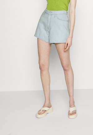 Persoon die lichtblauwe denimshorts draagt, een felgroene mouwloze top en crèmekleurige plateausandalen, staand op een effen achtergrond.