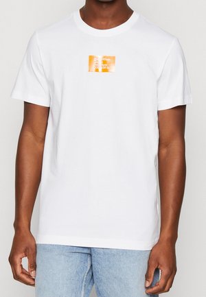Camiseta de algodón blanca con mangas cortas, que presenta un parche de logotipo cuadrado naranja en el pecho. Jeans de mezclilla azul claro visibles por debajo.