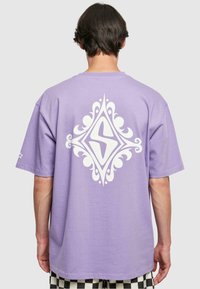 Purple cotton t-shirt with short sleeves featuring a large white decorative logo on the back, displayed against a neutral background.