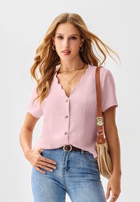 Lichtroze blouse met korte mouwen en een golvende halslijn, gouden knopen, gestructureerde stof, gecombineerd met blauwe denim en een beige handtas.