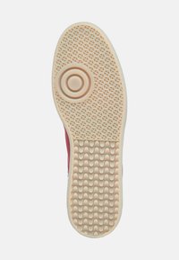 Gummisohle eines Sneakers mit einer strukturierten beige Oberfläche, die ein wellenförmiges Muster und ein kreisförmiges Rillendesign aufweist. Roter oberer Rand sichtbar.