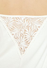 Top bianco con un pannello in pizzo a forma di V con un motivo foglia, bordato da delicati bordi ondulati, che offre un contrasto testurizzato con il tessuto.