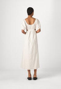 Lang, wit linnen jurk met een ronde rug, pofmouwen en uitsnijdingen aan de zijkanten. Heeft een gecollecteerde taille en een vloeiende uitstraling.