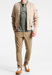 Veste bomber beige sur un polo vert, associée à un pantalon kaki et des baskets orange. Comprend des poches, des fermetures éclair et une coupe décontractée.