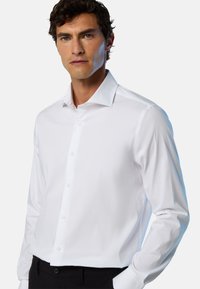 Weißes Langarmhemd aus glattem Stoff, mit spitzem Kragen, Knopfleiste und tailliertem Design. Kombiniert mit schwarzen Hosen.