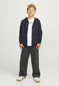 Temno modra zip-up kapuca s žepki čez belo grafično majico, v kombinaciji s temnimi hlačami in športnimi copati. Ohlapen dizajn s prijetno teksturo.