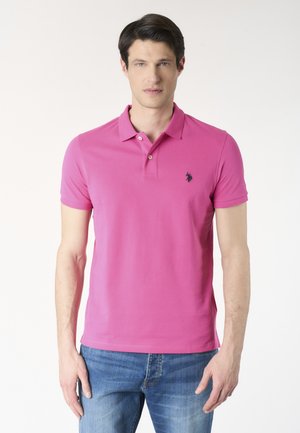 Uomo che indossa una polo rosa vivace con due bottoni e jeans blu, in piedi davanti a uno sfondo bianco semplice.