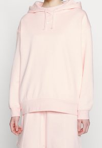 Sudadera con capucha de algodón rosa, con un bolsillo frontal y puños elásticos. Presenta un pequeño logo blanco en el pecho y un ajuste holgado.