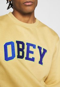 Beige sweatshirt featuring bold, blue "OBEY" text outlined in green. Crew neck design with a smooth texture and casual fit.