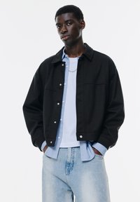 Jachetă neagră cu design cu guler, nasturi cu prindere argintie, suprapusă peste o cămașă cu dungi albastre deschise, asortată cu blugi din denim deschis.