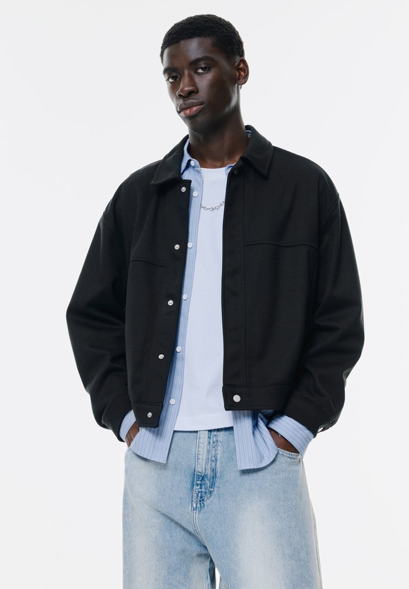 Jachetă neagră cu design cu guler, nasturi cu prindere argintie, suprapusă peste o cămașă cu dungi albastre deschise, asortată cu blugi din denim deschis.