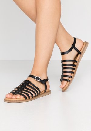 Pieds portant des sandales plates noires à brides avec boucle à la cheville et semelles couleur cuir sur un fond clair uni.