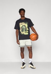 Melna T-krekls ar krāsainu grafiku, kopā ar baltām ventilējošām šortiem ar melniem svītrām, baltām zeķēm un basketbola apaviem. Tur basketbola bumbu.