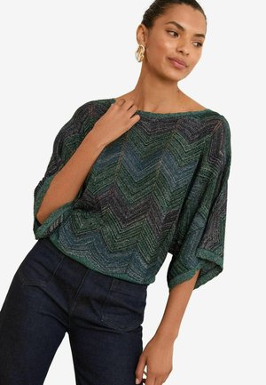 Grünes und navy blaues, mit Zickzackmuster gestricktes Oberteil mit weiten Ärmeln, runder Ausschnitt und strukturiertem Finish. Kombiniert mit dunklen Jeans.