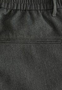 Tissu gris foncé avec une ceinture élastique et un détail de poche passepoilée horizontale, montrant la texture et les coutures de près.