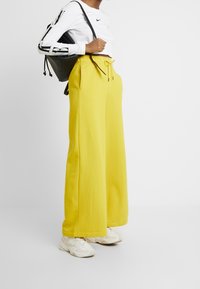 Gula vida byxor med fickor och dragsko, kombinerat med en långärmad vit Nike-tröja och vita sneakers. Svart ryggsäck.