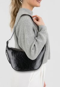 Borsa a spalla nera in pelle sintetica con forma curva, texture sottile e tracolla regolabile, indossata con un maglione grigio in maglia.