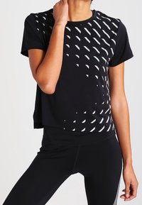 Musta urheilupaita, jossa on cropped-muotoilu ja valkoisella ylävasemmalla puolella oleva teksturoitu geometrinen kuvio. Yhdistetty mustiin leggings-housuihin.