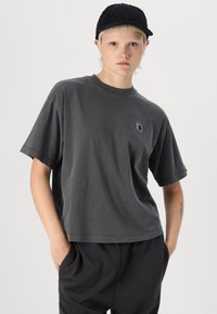 Gray cotton t-shirt with a crew neck, dropped shoulders, and a small logo patch. Worn with relaxed black pants and a black cap.