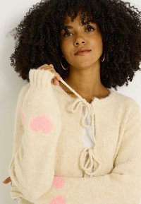 Cárdigan beige de punto con acabado texturizado, con motivos de corazones rosas en las mangas. Se ata en la parte delantera y tiene puños acanalados. Diseño suave y acogedor.