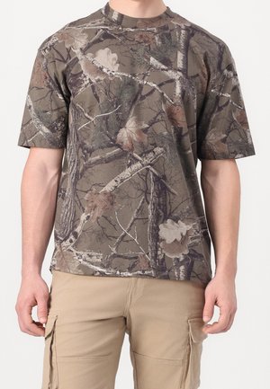 Korte mouwen overhemd met bruin-groen camouflagetak- en bladpatroon, gedragen met beige cargobroek.