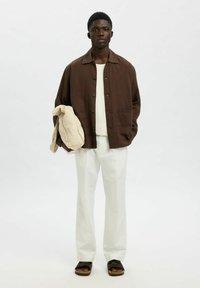 Brun knappad jacka över en vit texturerad skjorta, kombinerad med krämfärgade byxor och svarta sandaler; håller i en beige väska. Enkel design.