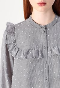 Blusa a scacchi bianca e nera con colletto arricciato, chiusura con bottoni e tessuto testurizzato con piccoli puntini in rilievo.