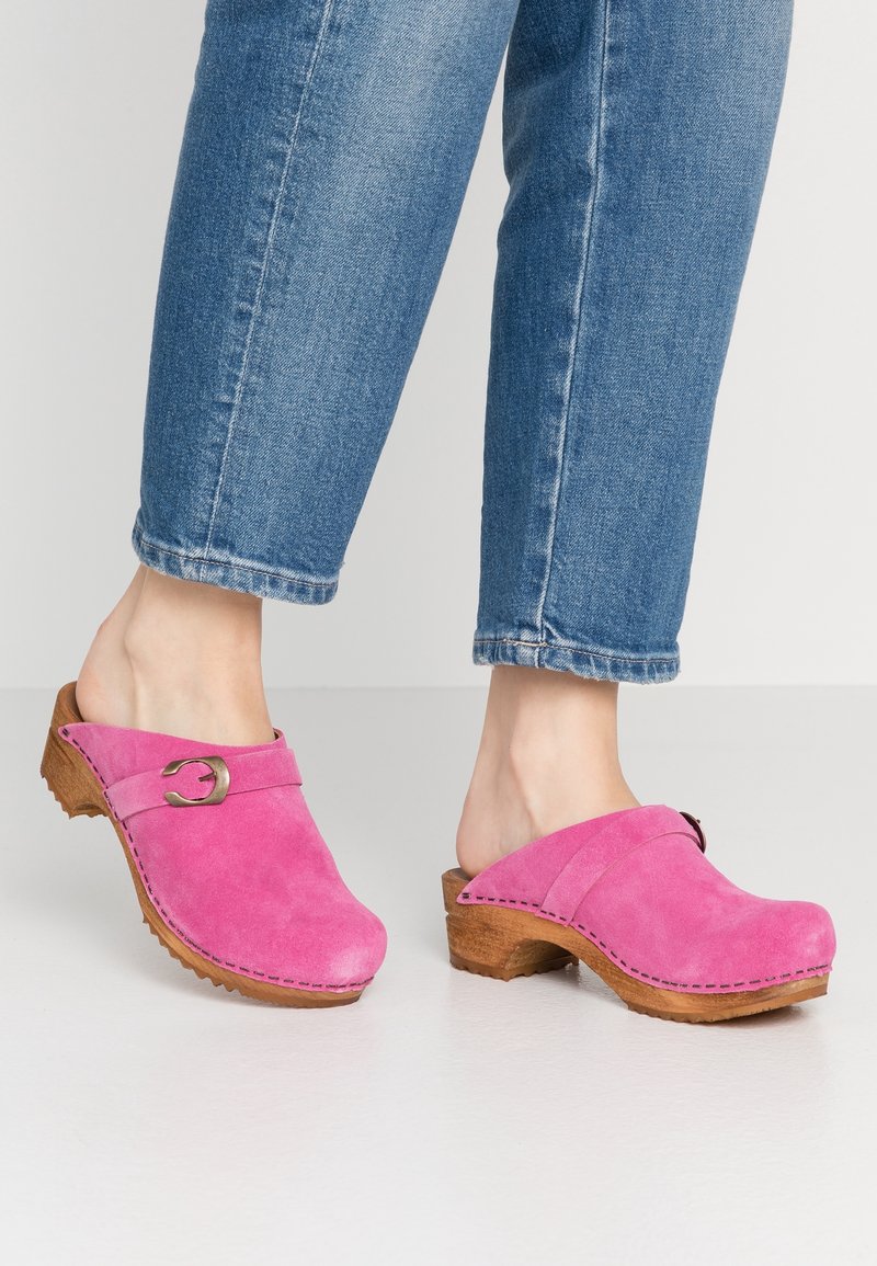 Personne portant un jean bleu et des sabots en daim rose vif avec semelles en bois et détail à boucle, debout sur un sol clair.