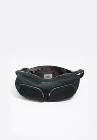 Ciemnozielona torba materiałowa z dwiema przednimi kieszeniami na zamek, o zaokrąglonym kształcie i miękkiej fakturze. Wewnątrz znajduje się beżowa podszewka z metką marki.