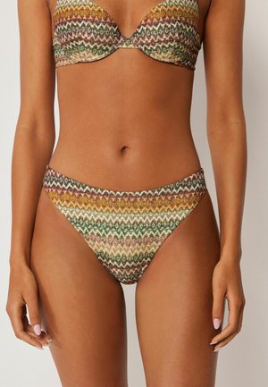 Kobieta nosząca bikini w wielokolorowy wzór zygzaka, z majtkami o wysokim kroju, na jasnym jednolitym tle.