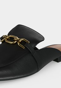 Μαύρο slip-on παπούτσι με υφή, μυτερή μύτη και λεπτομέρεια με χρυσή αλυσίδα στο επάνω μέρος, χαμηλό τακούνι ορατό δεξιά.