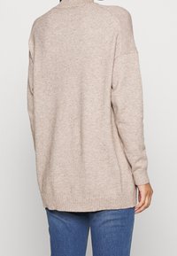 Helle beige Strickjacke mit hohem Halsausschnitt, überschnittenen Schultern, gerippten Bündchen und Saum. Lockere Passform mit einer strukturierten Stoffoberfläche.